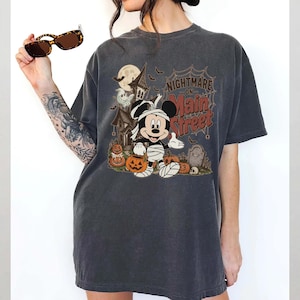 May include: Dark gray t-shirt featuring a Halloween-themed graphic. The design includes Mickey Mouse dressed as a mummy, pumpkins, a haunted house, and the text "Nightmare on Main Street".