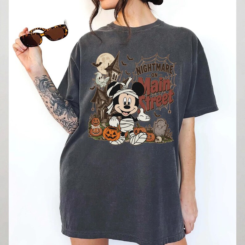 May include: Dark gray t-shirt featuring a Halloween-themed graphic. The design includes Mickey Mouse dressed as a mummy, pumpkins, a haunted house, and the text "Nightmare on Main Street".