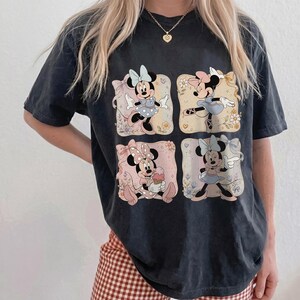 May include: Black t-shirt featuring four square illustrations of Minnie Mouse in various poses. Each image has a floral background and a pastel color scheme. The shirt is a relaxed fit.