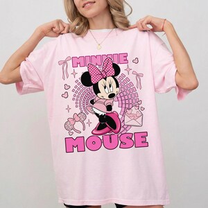 May include: Pale pink t-shirt with a Minnie Mouse design. The graphic shows Minnie Mouse in a pink dress and bow, with the words "MINNIE MOUSE" in pink. The shirt has short sleeves and a round neckline.