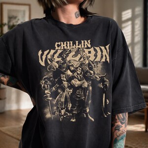 May include: A charcoal black short-sleeve t-shirt featuring a distressed beige graphic print of various animated villains. The text "CHILLIN Like a VILLAIN" is printed in a bold, stylized font above the group of characters on the front of the shirt.