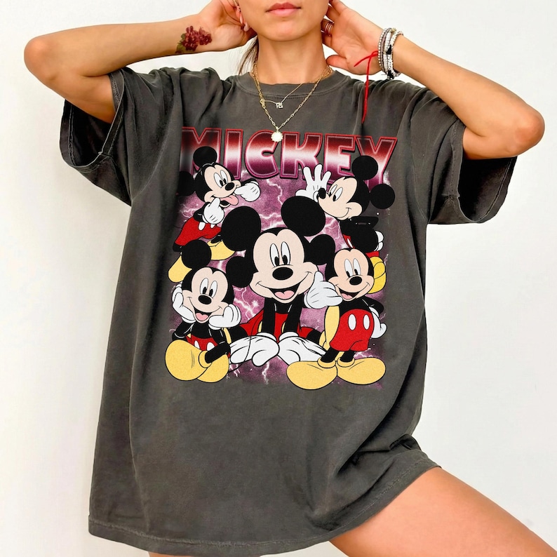 Puede incluir: Camiseta gris oscuro con un gr&aacute;fico de Mickey Mouse en varias poses. La palabra "MICKEY" est&aacute; impresa en rojo y blanco. La camiseta tiene un aspecto vintage y desgastado.
