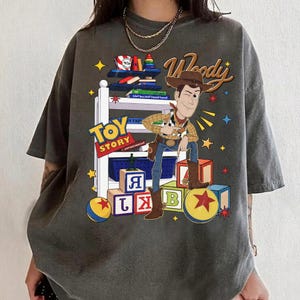 K&ouml;nnte beinhalten: Dunkelgraues T-Shirt mit einer Grafik von Woody aus Toy Story. Das Design enth&auml;lt die W&ouml;rter "Woody" und "Toy Story" sowie bunte Bl&ouml;cke, B&uuml;cher und Sterne. Woody wird in seiner Cowboy-Kleidung dargestellt.