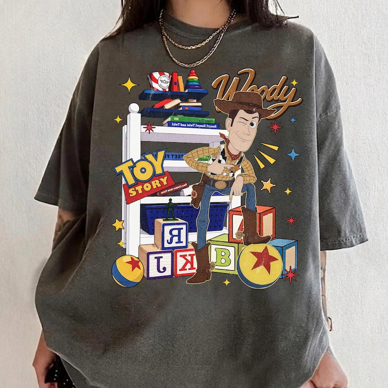 K&ouml;nnte beinhalten: Dunkelgraues T-Shirt mit einer Grafik von Woody aus Toy Story. Das Design enth&auml;lt die W&ouml;rter "Woody" und "Toy Story" sowie bunte Bl&ouml;cke, B&uuml;cher und Sterne. Woody wird in seiner Cowboy-Kleidung dargestellt.