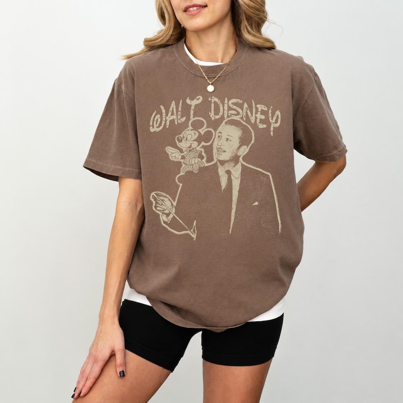 May include: Brown t-shirt featuring a vintage-style graphic of Walt Disney with Mickey Mouse and the text "Walt Disney". The shirt has a relaxed fit and short sleeves. The model is wearing black shorts and a white undershirt.