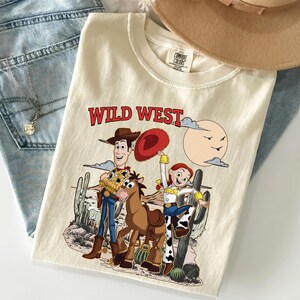 May include: Cream-colored t-shirt with a "Wild West" graphic featuring cartoon characters, a horse, and desert scenery. The shirt is folded with a hat and jeans in the background.