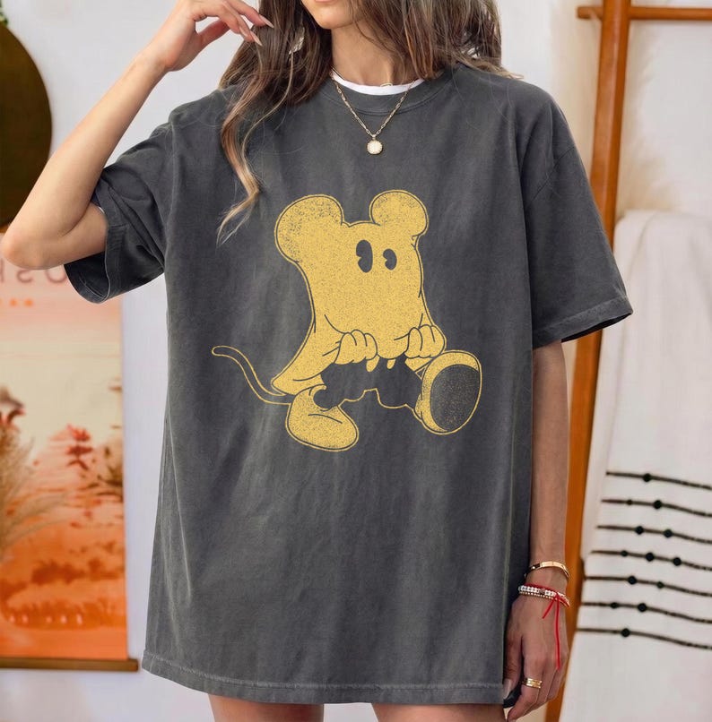 May include: Dark grey t-shirt featuring a yellow cartoon mouse design. The mouse is sitting with its hands clasped and has a long tail. The shirt has short sleeves and a relaxed fit, suitable for casual wear.