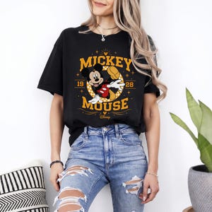 May include: Black t-shirt featuring a Mickey Mouse graphic. The design includes the words "Mickey Mouse" in gold lettering, along with the numbers "19" and "28". Mickey Mouse is depicted in his classic pose, wearing red shorts and white gloves.