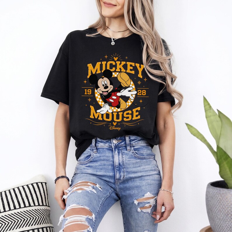 May include: Black t-shirt featuring a Mickey Mouse graphic. The design includes the words "Mickey Mouse" in gold lettering, along with the numbers "19" and "28". Mickey Mouse is depicted in his classic pose, wearing red shorts and white gloves.