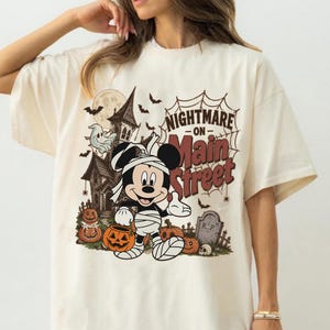 May include: Cream-colored t-shirt featuring a Halloween-themed graphic. The design includes a haunted house, bats, pumpkins, and a cartoon character dressed as a mummy. Text on the shirt reads "Nightmare on Main Street."