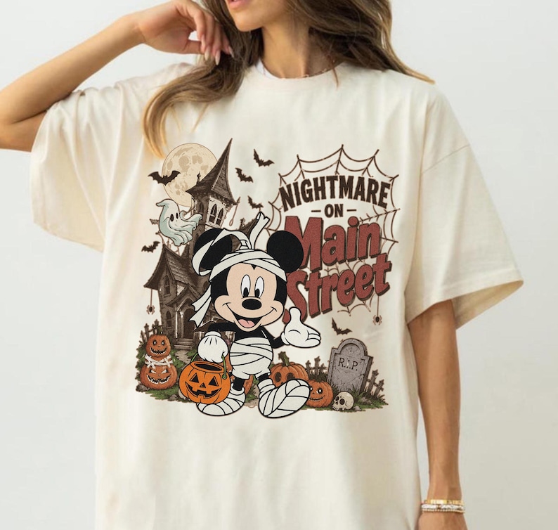 May include: Cream-colored t-shirt featuring a Halloween-themed graphic. The design includes a haunted house, bats, pumpkins, and a cartoon character dressed as a mummy. Text on the shirt reads "Nightmare on Main Street."