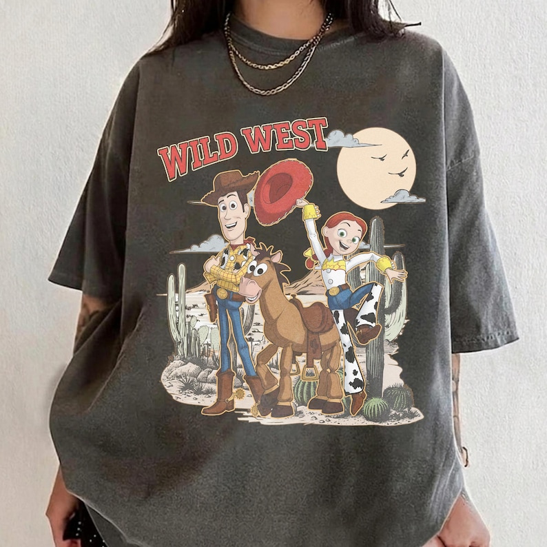 May include: Dark gray t-shirt featuring a Wild West graphic with cartoon characters, a horse, and cacti. The text "WILD WEST" is in red. The design includes a cowboy, cowgirl, and a horse in a desert landscape.