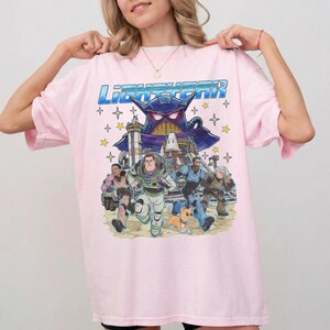 May include: Light pink t-shirt featuring a graphic of characters from the movie Lightyear. The design includes Buzz Lightyear, Zurg, and other characters, with the word "Lightyear" in blue. The shirt has a crew neck and short sleeves.