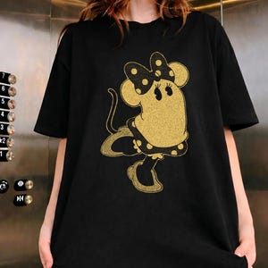 Pode incluir: T-shirt preto com um design de Minnie Mouse dourada com glitter. A personagem de desenho animado &eacute; retratada com um la&ccedil;o, vestido e sapatos de bolinhas. O design tem um aspeto vintage e texturizado, adicionando um toque de nostalgia &agrave; roupa.