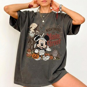 May include: Dark gray t-shirt featuring a Halloween-themed graphic. The design includes a cartoon mouse dressed as a mummy, pumpkins, a haunted house, and the text "Nightmare on Main Street."