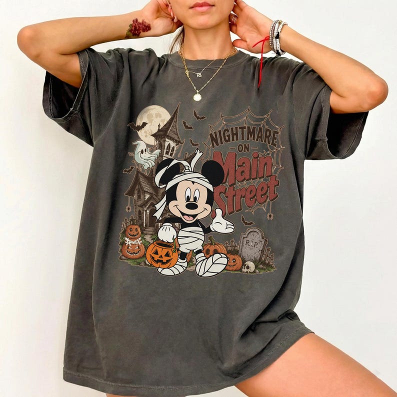 May include: Dark gray t-shirt featuring a Halloween-themed graphic. The design includes a cartoon mouse dressed as a mummy, pumpkins, a haunted house, and the text "Nightmare on Main Street."