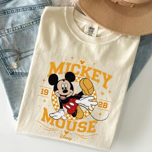 May include: Cream-colored t-shirt featuring a graphic of Mickey Mouse. The design includes the words "Mickey Mouse" in orange, along with the years "19" and "28". The Disney logo is at the bottom. The shirt is folded on top of blue jeans.