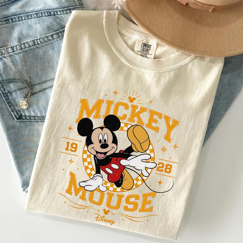 May include: Cream-colored t-shirt featuring a graphic of Mickey Mouse. The design includes the words "Mickey Mouse" in orange, along with the years "19" and "28". The Disney logo is at the bottom. The shirt is folded on top of blue jeans.