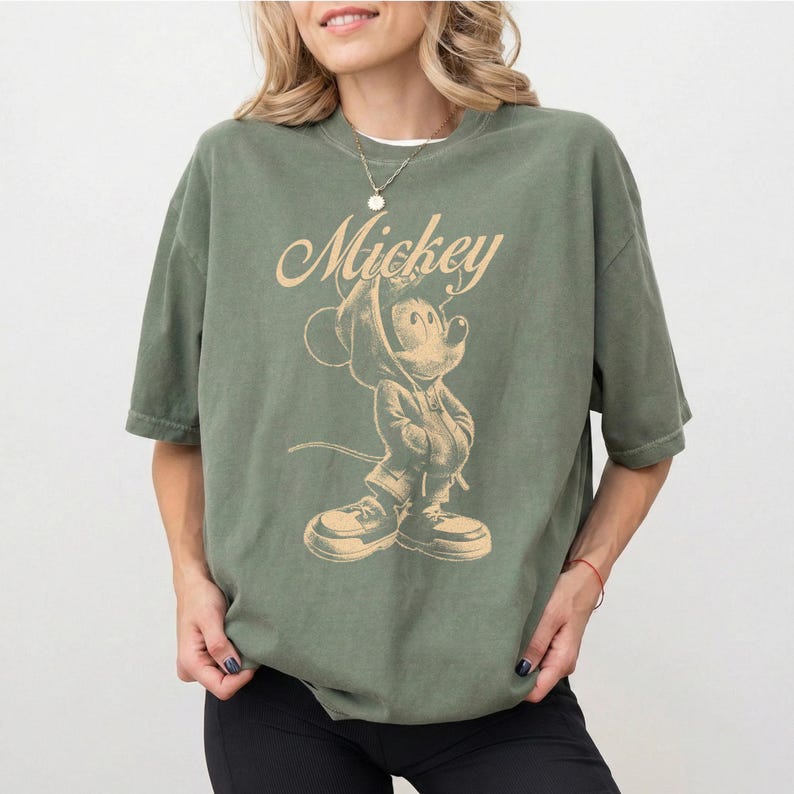 Puede incluir: Una camiseta de manga corta color verde oliva con un estampado gr&aacute;fico en beige de un rat&oacute;n de dibujos animados con sudadera con capucha y zapatillas. La palabra Mickey est&aacute; impresa en letra cursiva sobre la ilustraci&oacute;n del personaje.