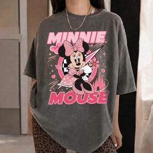 May include: Gray t-shirt featuring a graphic of Minnie Mouse in a pink dress, bow, and holding a flower. The words "MINNIE MOUSE" are in pink above and below the character. A pink and white circle and a pink castle are also in the design.