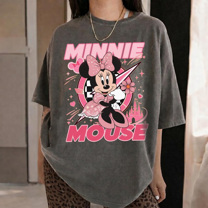 May include: Gray t-shirt featuring a graphic of Minnie Mouse in a pink dress, bow, and holding a flower. The words "MINNIE MOUSE" are in pink above and below the character. A pink and white circle and a pink castle are also in the design.
