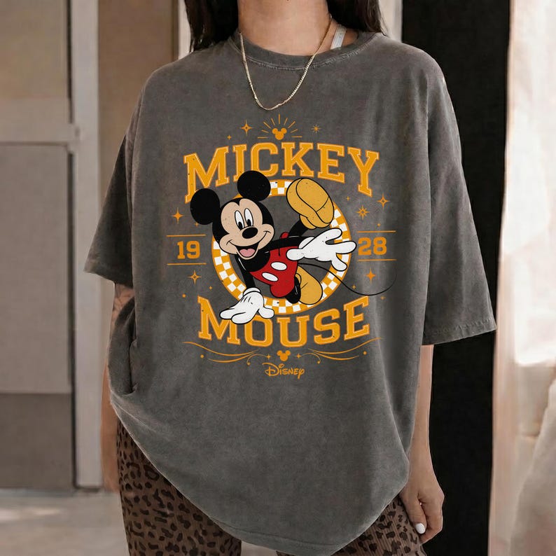May include: A gray t-shirt featuring a graphic of Mickey Mouse in a dynamic pose. The design includes the words "Mickey Mouse" in yellow, along with the year "1928" and the Disney logo. The shirt has short sleeves.