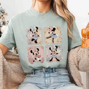 May include: A light green t-shirt featuring four square illustrations of Minnie Mouse in various poses. Each illustration has a different background color and floral accents. Minnie Mouse is wearing different outfits and bows in each illustration.