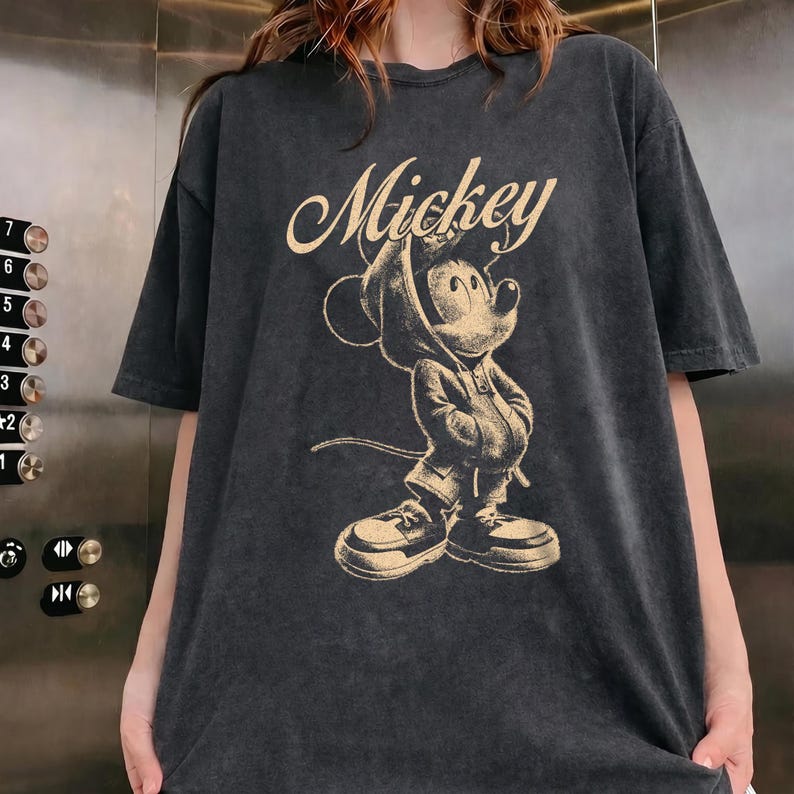 Puede incluir: Una camiseta de manga corta gris carb&oacute;n con un estampado gr&aacute;fico en beige de Mickey Mouse vistiendo una sudadera con capucha y zapatillas. La palabra Mickey est&aacute; impresa en una fuente cursiva sobre la ilustraci&oacute;n del personaje.