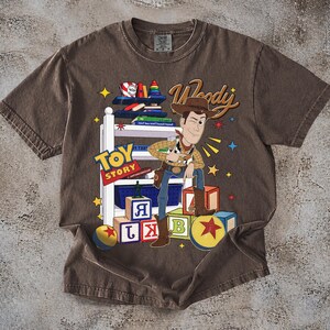 K&ouml;nnte beinhalten: Braunes T-Shirt mit einer Grafik von Woody aus Toy Story. Das Design enth&auml;lt die W&ouml;rter "Toy Story" und "Woody" sowie bunte Bl&ouml;cke und Sterne. Das T-Shirt hat einen Vintage-Look.