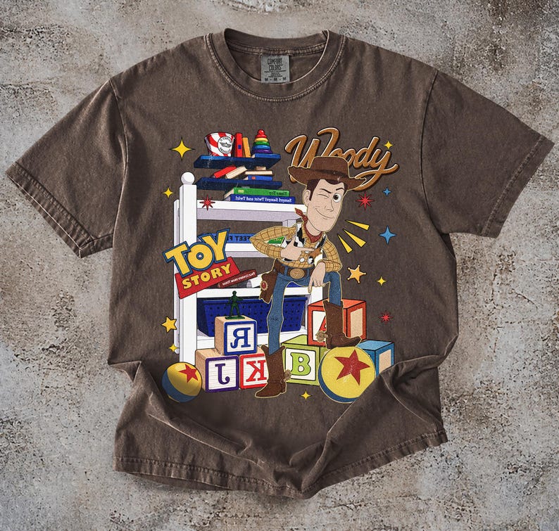 K&ouml;nnte beinhalten: Braunes T-Shirt mit einer Grafik von Woody aus Toy Story. Das Design enth&auml;lt die W&ouml;rter "Toy Story" und "Woody" sowie bunte Bl&ouml;cke und Sterne. Das T-Shirt hat einen Vintage-Look.