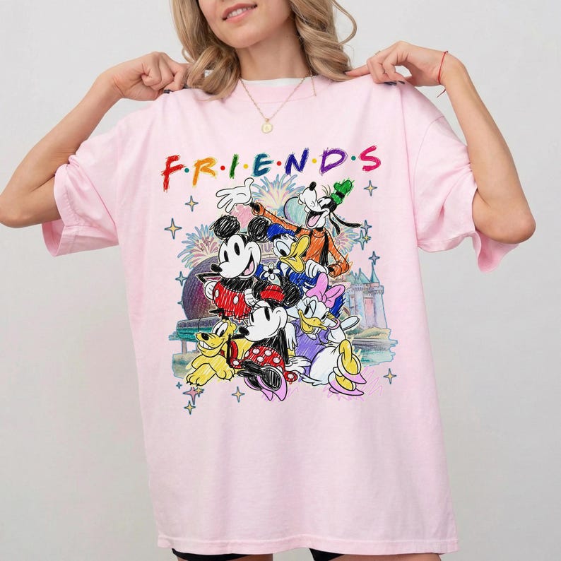May include: A light pink t-shirt featuring the word "FRIENDS" in colorful, stylized lettering. Below the text is a graphic of Disney characters including Mickey Mouse, Minnie Mouse, Donald Duck, Goofy, and Daisy Duck. The design includes fireworks and castle imagery.