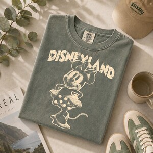 May include: A sage green t-shirt featuring a distressed "Disneyland" graphic and a Minnie Mouse illustration. The shirt is surrounded by a beige cap, a mug, and green and beige sneakers.
