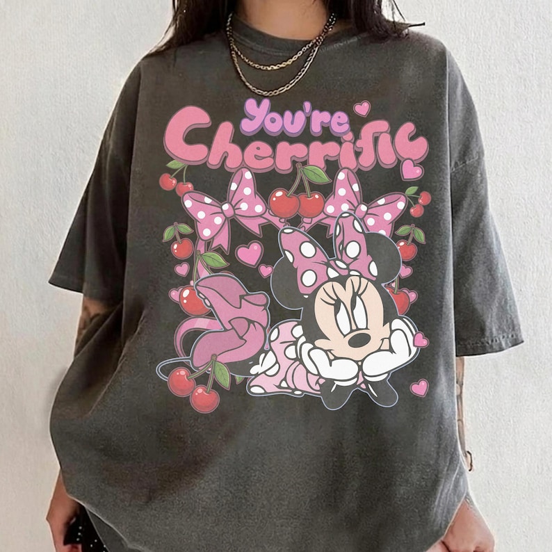 Pode incluir: T-shirt cinzento escuro com Minnie Mouse e o texto "You're Cherrific". O design inclui la&ccedil;os cor-de-rosa, cerejas e cora&ccedil;&otilde;es. A camisola tem um aspeto vintage e desgastado.