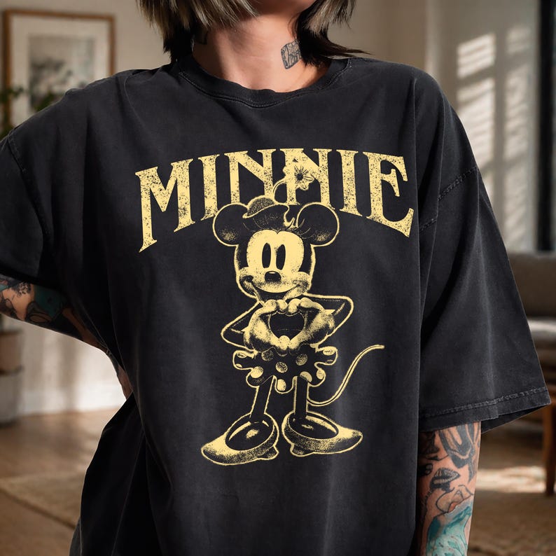 May include: Black t-shirt featuring a distressed yellow graphic of Minnie Mouse. The word "MINNIE" is arched above the character. Minnie is depicted with a heart-shaped gesture. The shirt has a vintage, worn look.