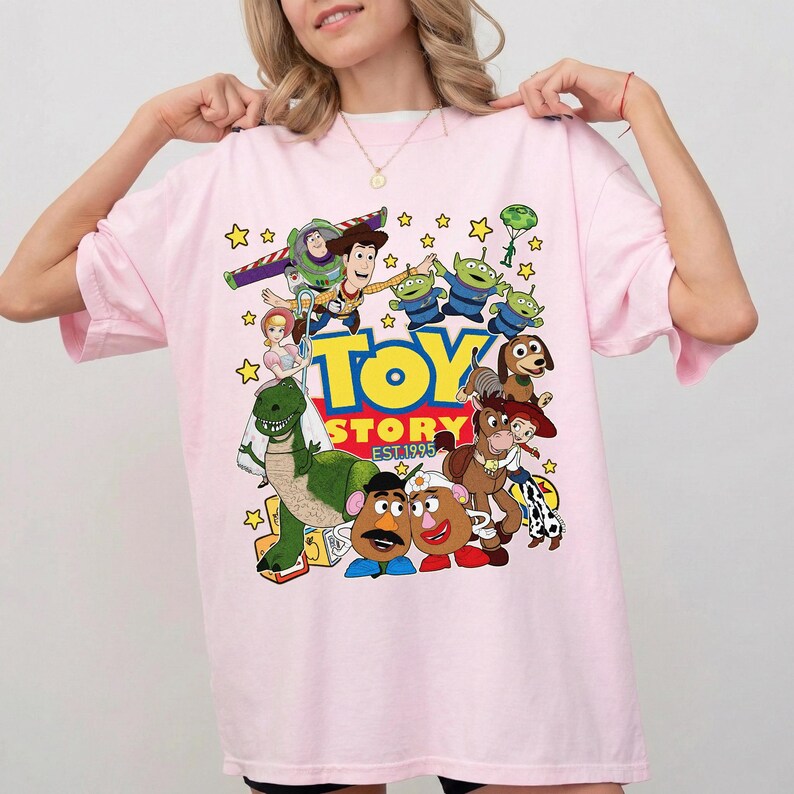 Pu&ograve; includere: T-shirt rosa chiaro con una grafica colorata dei personaggi del film d'animazione "Toy Story". Il design include Woody, Buzz Lightyear, gli alieni e altri personaggi, con la scritta "TOY STORY EST. 1995."