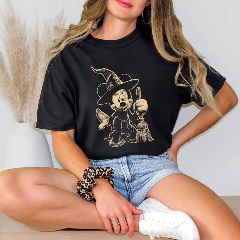 Puede incluir: Camiseta negra con un dise&ntilde;o en tonos sepia de Minnie Mouse vestida de bruja, sosteniendo una escoba. El dise&ntilde;o incluye un sombrero de bruja y una Minnie sonriente. La camiseta tiene un corte holgado y extragrande.