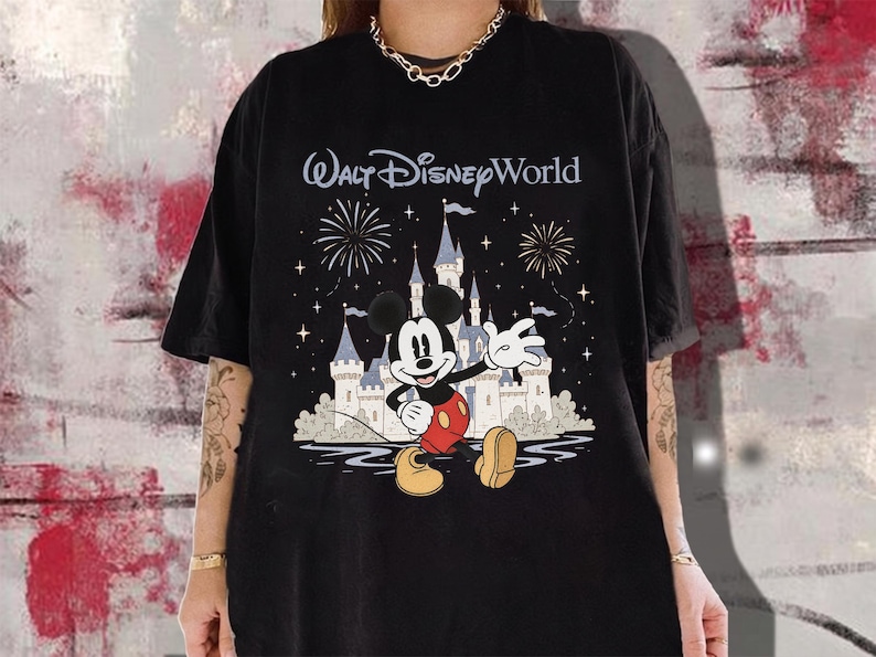 May include: Black t-shirt featuring Mickey Mouse waving in front of Cinderella's castle with fireworks and the text "Walt Disney World". The design is in a cartoon style with a blue, white, red, and yellow color palette.