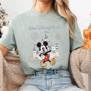 May include: A light green t-shirt featuring a graphic of Mickey Mouse waving in front of a castle with fireworks and the text "Walt Disney World". The shirt has short sleeves and a crew neck.