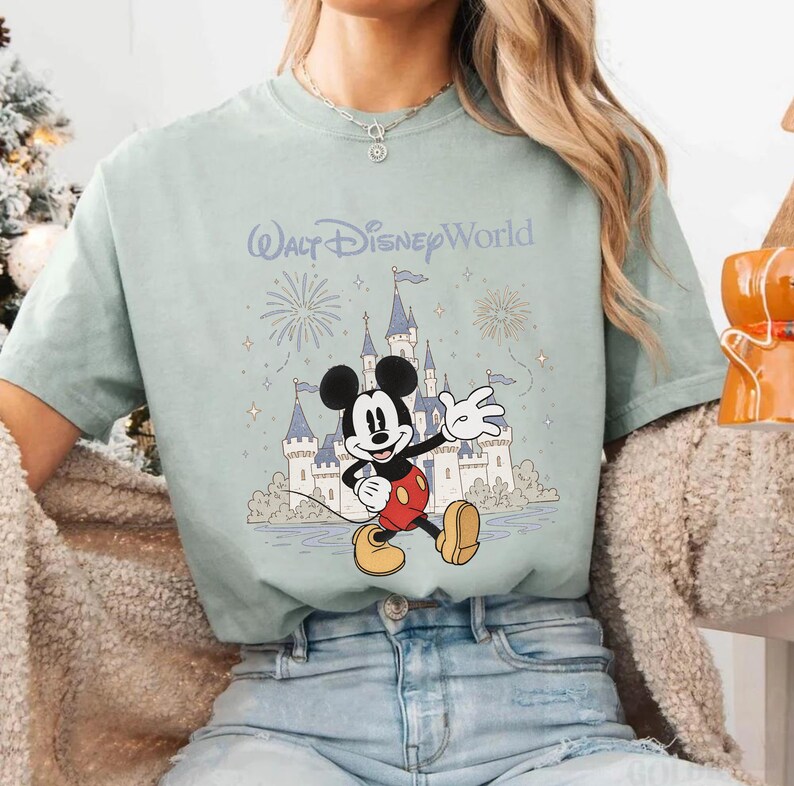 May include: A light green t-shirt featuring a graphic of Mickey Mouse waving in front of a castle with fireworks and the text "Walt Disney World". The shirt has short sleeves and a crew neck.