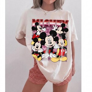 Puede incluir: Camiseta color crema con un gr&aacute;fico de Mickey Mouse en varias poses. La palabra "MICKEY" est&aacute; impresa en una fuente degradada roja y morada en la parte superior. La camiseta se combina con pantalones cortos a cuadros rojos y blancos.