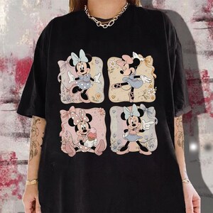 May include: Black t-shirt featuring four panels with Minnie Mouse illustrations. Each panel showcases Minnie in different poses, wearing dresses and bows. The background of each panel has floral and pastel elements.