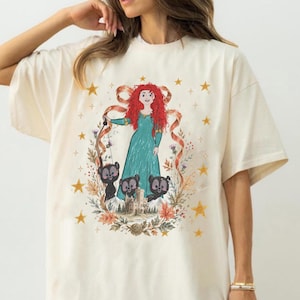 May include: Cream-colored t-shirt featuring a cartoon illustration of a character with red hair and a teal dress, surrounded by bears, a castle, and stars. The design is framed by a floral wreath.