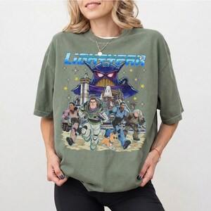 May include: Olive green t-shirt featuring a graphic from the movie Lightyear. The design includes the movie title, Buzz Lightyear, and other characters running from a large purple villain. The shirt has a relaxed fit.