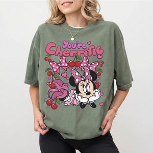 Pode incluir: Camiseta verde-oliva com um gr&aacute;fico da Minnie Mouse, la&ccedil;os rosa, cerejas e o texto "You're Cherrific". O design inclui cora&ccedil;&otilde;es e um padr&atilde;o de bolinhas. A camisa tem mangas curtas.