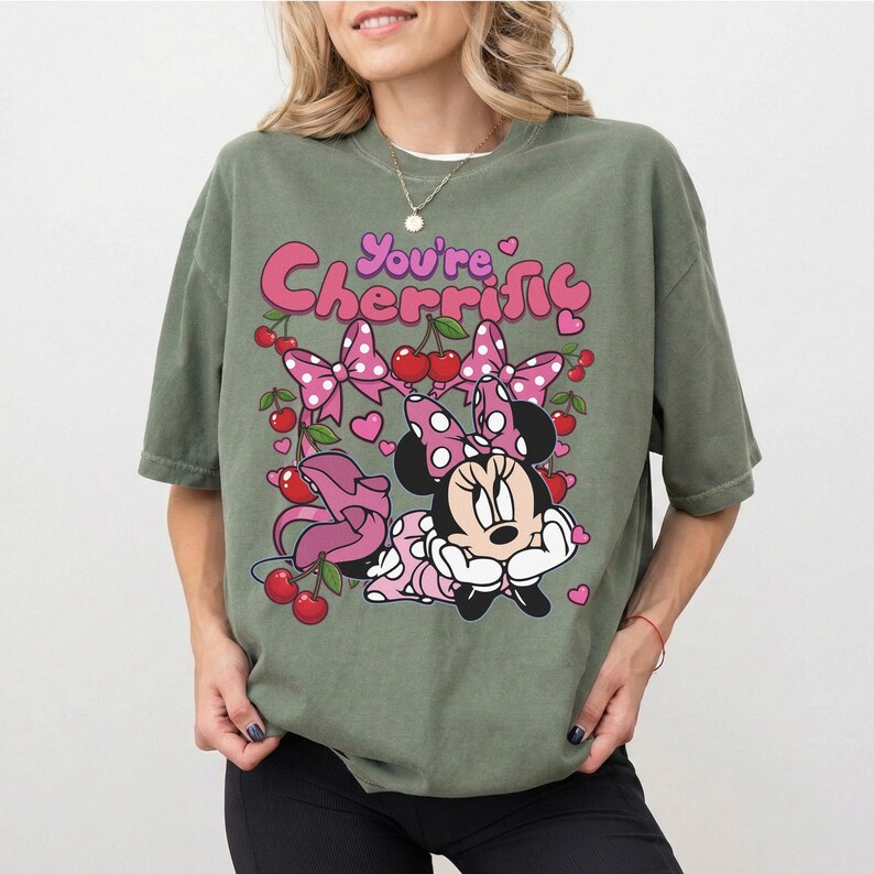 Pode incluir: Camiseta verde-oliva com um gr&aacute;fico da Minnie Mouse, la&ccedil;os rosa, cerejas e o texto "You're Cherrific". O design inclui cora&ccedil;&otilde;es e um padr&atilde;o de bolinhas. A camisa tem mangas curtas.
