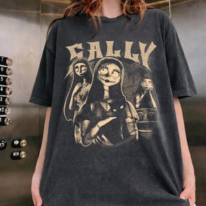 May include: A charcoal gray short-sleeve t-shirt featuring a distressed graphic print of the character Sally from The Nightmare Before Christmas. The design includes the text SALLY in a large, stylized font above three illustrations of the character.