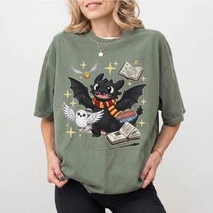 May include: Sage green t-shirt featuring a cartoon dragon with a red and yellow scarf, a white owl, and open books. The design includes a golden snitch and stars, evoking a fantasy theme.