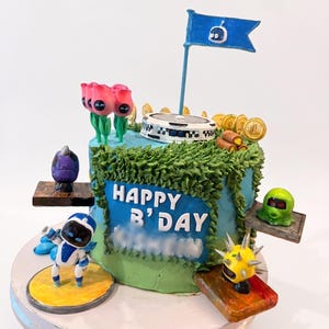 May include: A round blue cake decorated with green frosting vines, featuring the text HAPPY B'DAY. The cake is topped with a blue flag, gold coins, and small figures including a white and blue robot, a purple creature, and a yellow spiked character.