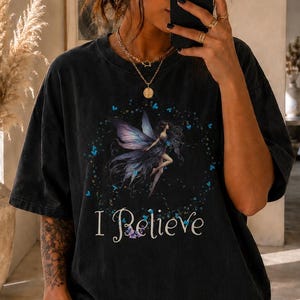 May include: A black short-sleeved t-shirt featuring a graphic print of a fairy with purple wings surrounded by small blue butterflies and the text I Believe in white script lettering. The shirt has a relaxed fit and a faded, vintage-style wash.