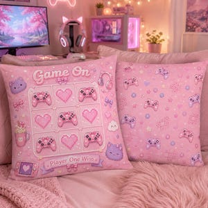 May include: Two square throw pillows in light pink featuring a gaming theme. One pillow displays a tic-tac-toe grid with game controllers and hearts, the text "Game On" and "Player One Wins." The other pillow has a repeating pattern of controllers.